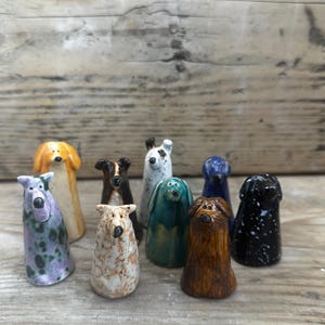 May include: A collection of eight small, hand-sculpted ceramic dog figurines in various colours and patterns, including solid brown, speckled black, teal, purple, white, and tan, each featuring simple painted facial details and upright postures.