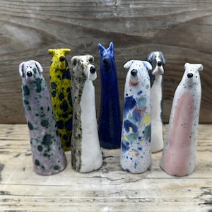 May include: Seven tall, slender ceramic dog figurines with distinct glazes. The collection includes a purple spotted dog, a yellow and blue spotted dog, a white and brown speckled dog, a solid blue dog, a white dog with blue and yellow splashes, a grey and white dog, and a white dog with a pink patch.