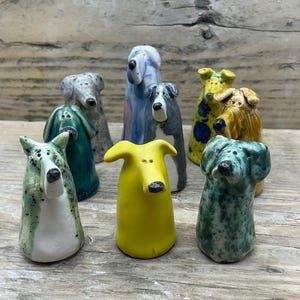 May include: A collection of eight small, hand-sculpted ceramic dog figurines in various colours and patterns. The group includes solid yellow, teal, and multi-coloured glazed pieces with spotted, striped, and textured finishes, each featuring a black nose.