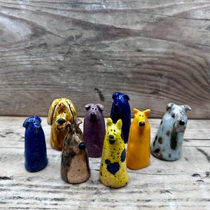 May include: A collection of eight small, hand-sculpted ceramic dog figurines in various colours and patterns, including solid blue, yellow, and purple, as well as spotted and multi-coloured designs, each featuring a simple, upright, conical shape.