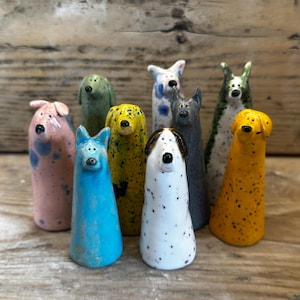 May include: A collection of eight small, cone-shaped ceramic dog figurines in various colours including pink, blue, yellow, white with black spots, and grey. Each figurine features a simple painted face with eyes and a nose, standing upright.