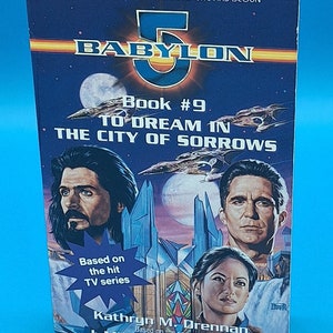 May include: Book cover for "Babylon 5: To Dream in the City of Sorrows", book number 9 in the series. The cover features a futuristic cityscape with three characters, two men and one woman, looking out at the viewer. The text "Based on the hit TV series" is visible in the lower left corner. The authors are Kathryn M. Drennan and J. Michael Straczynski.