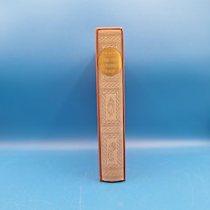 May include: A hardcover book spine titled The Old Curiosity Shop by Dickens, featuring a tan background with gold embossed lettering and intricate decorative borders, including two small illustrations of figures in rectangular frames.