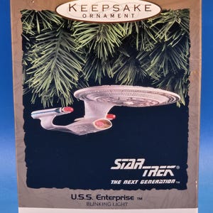 May include: A Hallmark Keepsake Ornament box featuring the U.S.S. Enterprise from Star Trek: The Next Generation. The box displays the starship with red and white accents, the Star Trek logo, and text reading U.S.S. Enterprise Blinking Light Magic.