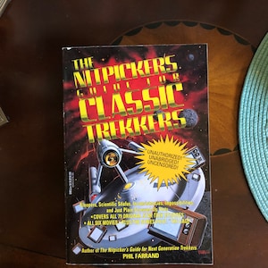 May include: A book cover with a red, orange, and black background featuring a spaceship and the text "The Nitpickers Guide for Classic Trekkers". The book cover also includes the text "Unauthorized! Unabridged! Uncensored!" and "Covers all 79 original Star Trek episodes. All six movies plus the series pilot "The Cage"."