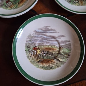 May include: Set of decorative plates with a hunting scene design. Each plate features a white center with a detailed illustration of riders on horseback, dogs, and a landscape. The plates have a green border.