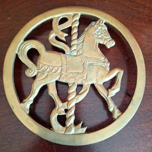 May include: A gold-colored metal carousel horse within a circular frame. The horse is detailed with a saddle and mane, and is attached to a central pole. The design is a decorative piece, possibly a trivet or wall hanging.