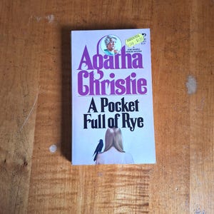 May include: A paperback book titled A Pocket Full of Rye by Agatha Christie. The cover features a purple title, a small circular portrait of a woman, and an illustration of a person with long blonde hair and a black bird perched on their shoulder.