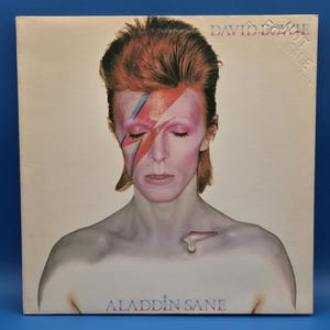 May include: A square vinyl record cover featuring a portrait of David Bowie with a red and blue lightning bolt painted across the face. The text DAVID BOWIE appears at the top right, and ALADDIN SANE is printed in blue at the bottom center.