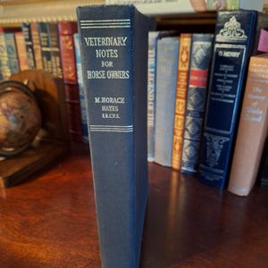 May include: A navy blue book titled "Veterinary Notes for Horse Owners" by M. Horace Hayes. The title and author's name are in white lettering. The book is standing upright among other books on a shelf.
