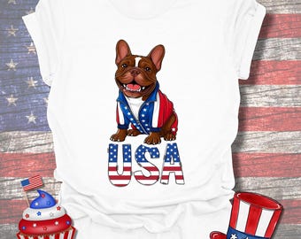 Puede incluir: Camiseta blanca con un bulldog francés de dibujos animados que lleva una chaqueta roja, blanca y azul. La palabra "USA" está impresa en una fuente patriótica. También se ven una magdalena y un sombrero con colores patrióticos.