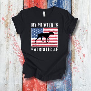May include: Black t-shirt with a patriotic design. The design features a silhouette of a dog, an American flag, and the text "MY POINTER IS PATRIOTIC AF" in white. The shirt is made of a soft material.