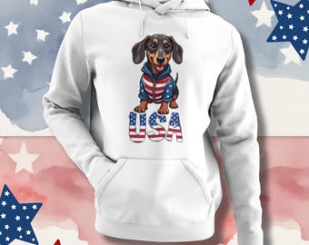 Könnte beinhalten: Weißer Kapuzenpullover mit einem Dackel, der eine Jacke in den Farben der amerikanischen Flagge trägt, mit dem Wort "USA". Das Design ist eine spielerische Feier des amerikanischen Stolzes, für Hundeliebhaber und patriotische Veranstaltungen.