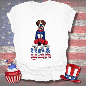 May include: White t-shirt with a seated dog wearing a jacket in the colours of the American flag. The word "USA" is printed below the dog in a matching design. A cupcake and a hat with patriotic colours are also visible.