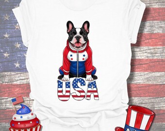 Puede incluir: Camiseta blanca con un bulldog francés que lleva una chaqueta roja, blanca y azul. El perro está sentado en la palabra "USA" en una fuente patriótica. También se ve una magdalena y un sombrero con estrellas y rayas.