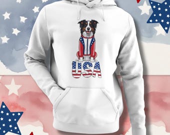 Könnte beinhalten: Weißer Kapuzenpullover mit einer Hundeillustration, die eine rot-weiß-blaue Jacke trägt. Das Wort "USA" ist in einer patriotischen Schriftart darunter gedruckt. Der Hintergrund hat einen Aquarelleffekt mit Sternen.