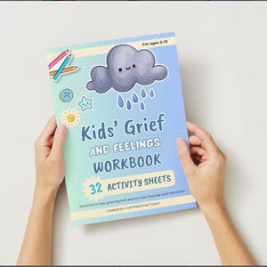 Puede incluir: Un cuaderno de trabajo titulado "Kids' Grief and Feelings Workbook" con una cubierta degradada azul y verde. La cubierta presenta una nube de dibujos animados con gotas de lluvia, una estrella, un sol y una cara triste. El cuaderno de trabajo está sostenido por dos manos.