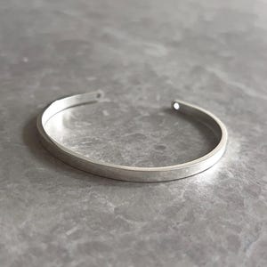 May include: A minimalist silver cuff bracelet. The open-ended bracelet has a flat, rectangular profile and a polished finish. The jewelry is displayed on a gray, marble-like surface. The bracelet is a simple, elegant accessory.