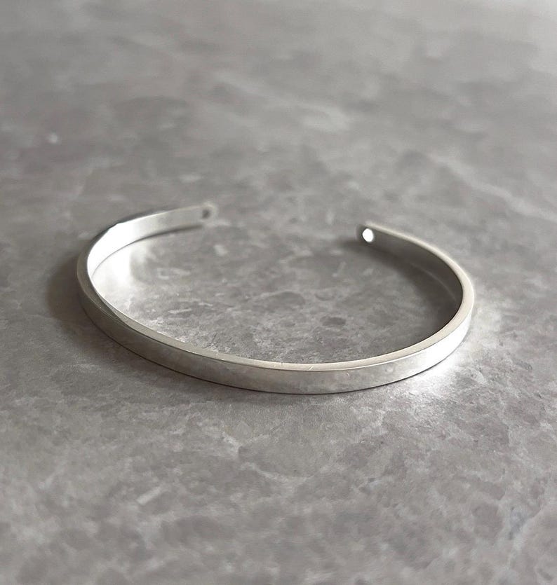May include: A minimalist silver cuff bracelet. The open-ended bracelet has a flat, rectangular profile and a polished finish. The jewelry is displayed on a gray, marble-like surface. The bracelet is a simple, elegant accessory.