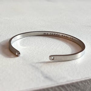May include: A silver-colored cuff bracelet with the engraved text "my teddy bear <3". The open-ended bracelet is displayed on a light-colored surface. The bracelet is a simple, elegant design.