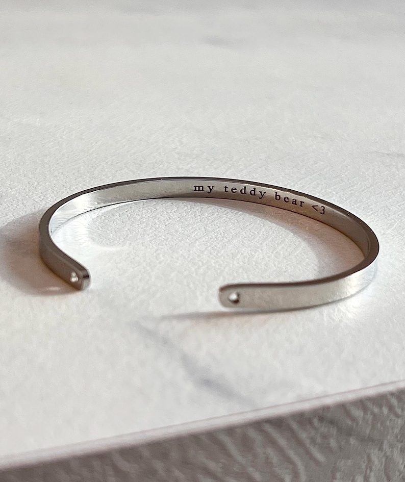 May include: A silver-colored cuff bracelet with the engraved text "my teddy bear <3". The open-ended bracelet is displayed on a light-colored surface. The bracelet is a simple, elegant design.