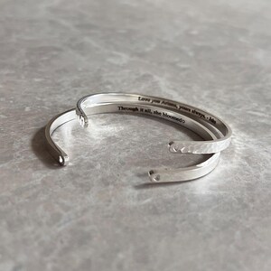 May include: Three silver-colored open cuff bracelets. The top bracelet has the inscription "Love you Ariana, yours always. - Jake" and "Through it all, she bloomed". The middle bracelet has a hammered texture.