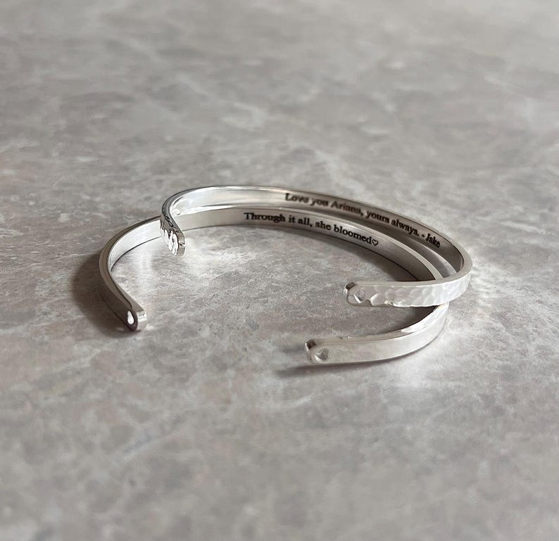 May include: Three silver-colored open cuff bracelets. The top bracelet has the inscription "Love you Ariana, yours always. - Jake" and "Through it all, she bloomed". The middle bracelet has a hammered texture.