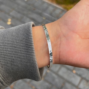 May include: A silver-colored bangle bracelet with a hammered texture. The bracelet is worn on a wrist, and the background is blurred. The bracelet appears to be made of metal and reflects light.