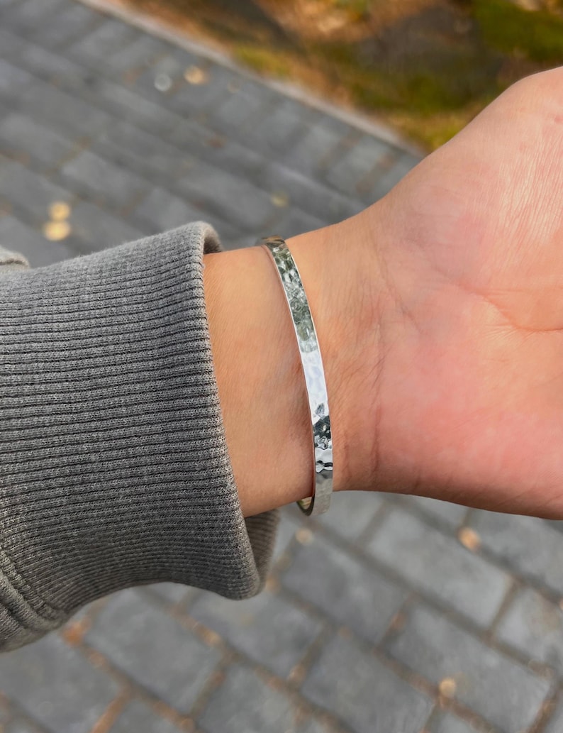 May include: A silver-colored bangle bracelet with a hammered texture. The bracelet is worn on a wrist, and the background is blurred. The bracelet appears to be made of metal and reflects light.