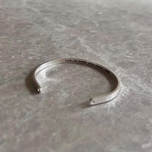 May include: A silver cuff bracelet with the engraved text "Love you Ariana, yours always - Jake". The bracelet is open-ended and rests on a gray marble surface.