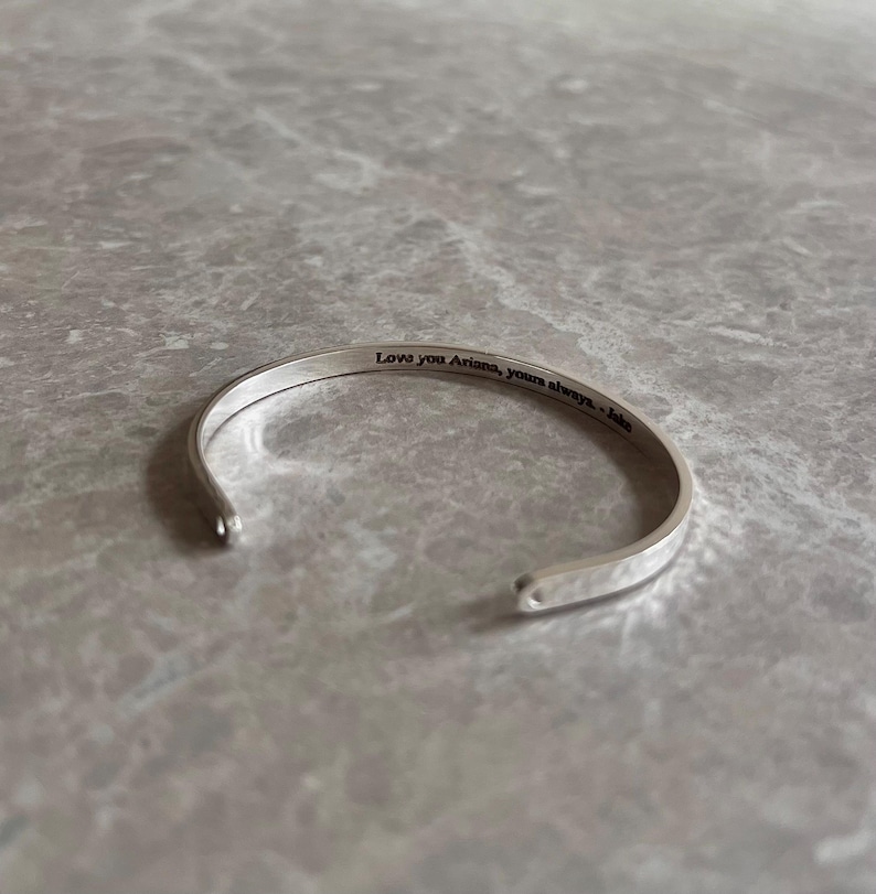 May include: A silver cuff bracelet with the engraved text "Love you Ariana, yours always - Jake". The bracelet is open-ended and rests on a gray marble surface.