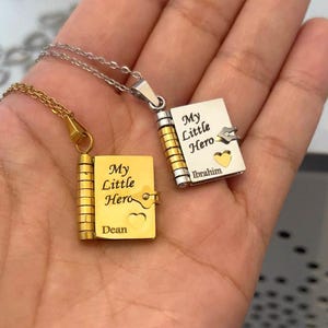 May include: Two pendant necklaces, each with a miniature book design. One is gold-toned, the other silver-toned. Both books have the words "My Little Hero" engraved, along with a name and a heart symbol. The necklaces are displayed on a hand.