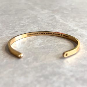 Op de afbeelding: Een goudkleurige metalen armband met een open ontwerp. De binnenzijde van de armband is voorzien van de gegraveerde zwarte tekst: My Love For You Is Infinite, Forever & Always. De armband heeft afgeronde uiteinden en een gladde, gepolijste afwerking.