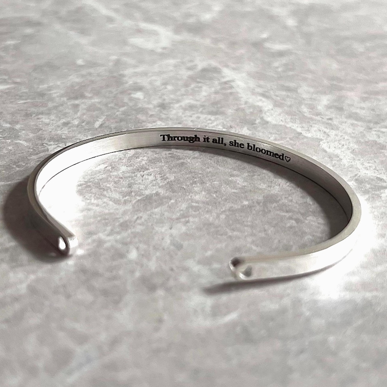 May include: A silver-colored cuff bracelet with the engraved text "Through it all, she bloomed" and a heart symbol. The open-ended bracelet is displayed on a light gray, textured surface.