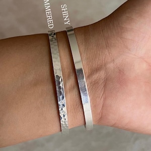 May include: Two silver bracelets are shown on a wrist. One is hammered, the other is shiny. The hammered bracelet has a textured surface, while the shiny bracelet is smooth. The bracelets are simple and elegant.