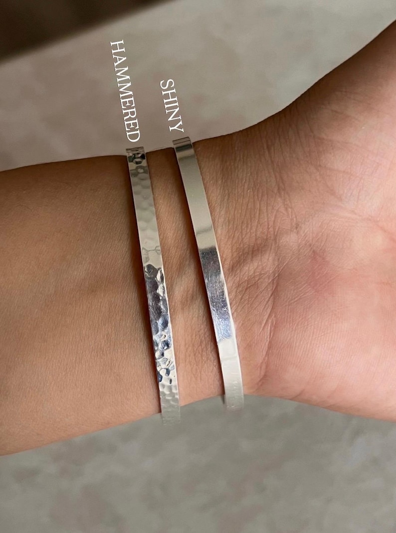 May include: Two silver bracelets are shown on a wrist. One is hammered, the other is shiny. The hammered bracelet has a textured surface, while the shiny bracelet is smooth. The bracelets are simple and elegant.