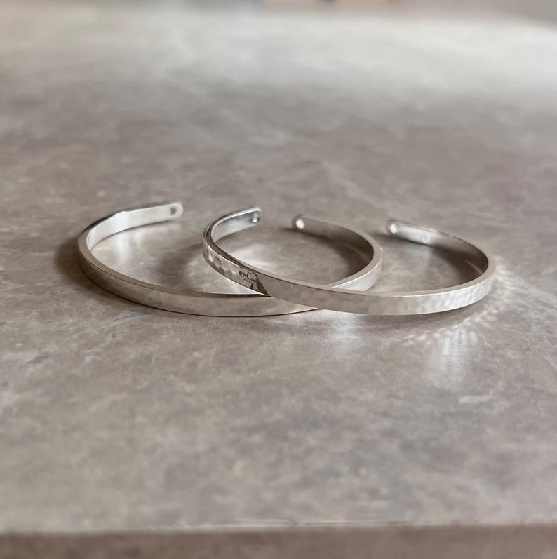 May include: Three silver-colored cuff bracelets. Two have a hammered texture, and one is smooth. The bracelets are open-ended and rest on a light gray surface. The bracelets are a simple, minimalist design.