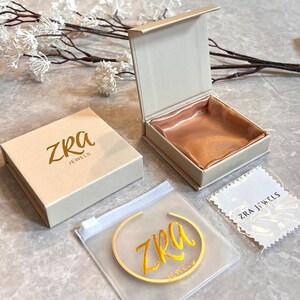 May include: A jewelry presentation set featuring a beige box with the gold text "ZRA JEWELS", an open box with a peach satin interior, a clear plastic bag with a gold bracelet, and a white cleaning cloth.
