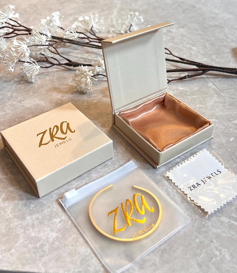 May include: A jewelry presentation set featuring a beige box with the gold text "ZRA JEWELS", an open box with a peach satin interior, a clear plastic bag with a gold bracelet, and a white cleaning cloth.