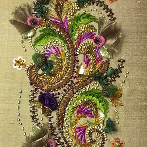 Puede incluir: Primer plano de un diseño de bordado detallado con un patrón en espiral de cuentas y lentejuelas doradas, moradas, verdes y marrones. El diseño está sobre un fondo de tela beige.