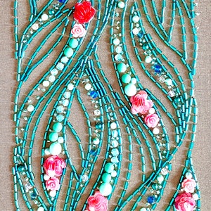 May include: A close-up of a beaded artwork featuring a pattern of turquoise and pink beads arranged in a floral design. The beads are sewn onto a beige fabric background.