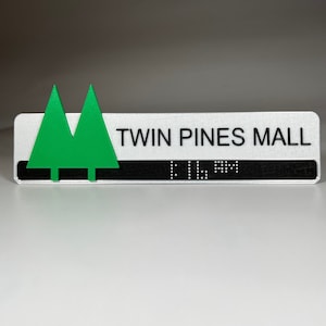May include: A white sign with black text that reads "Twin Pines Mall". The sign has a green, 3D, twin pine tree icon on the left side. The sign also has braille text below the mall name.