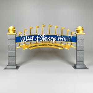 Puede incluir: Una réplica en miniatura del letrero de entrada de Walt Disney World. El letrero presenta las palabras "Walt Disney World" en escritura blanca sobre una pancarta azul, con detalles dorados y la frase "THE MOST MAGICAL PLACE ON EARTH". La estructura incluye pilares grises y adornos dorados.