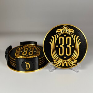 May include: A set of four black coasters with a gold design featuring the number "33" and a stylized floral motif. The coasters are stacked and one is on a stand.