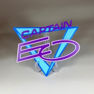 Puede incluir: Un letrero impreso en 3D de color morado y azul con el texto "CAPTAIN" y un logotipo estilizado "E" y "D".
