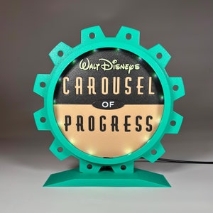Op de afbeelding: Een tealblauw tandwielvormig bord met een rond, verlicht display waarop "Walt Disney's Carousel of Progress" staat.