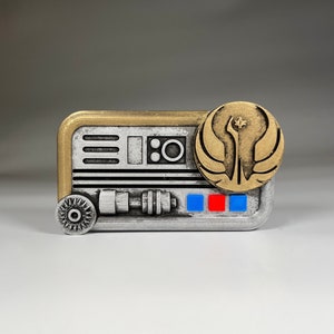 May include: A silver and gold metal pin with a Star Wars Rebel Alliance symbol and a small, detailed, silver and black design. The pin has a red and blue stripe design.