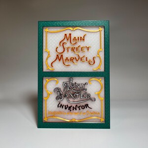 May include: A green sign with gold trim and the text "Main Street Marvels" and "Tony Baxter Inventor Imagination is at the heart of our creations."