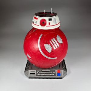 May include: A red and white Star Wars BB-8 droid toy with a silver base. The droid is on a stand with a silver base and a red and blue button.