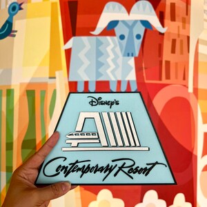 May include: A blue and white triangular sign with black text that reads "Disney's Contemporary Resort". The sign features a stylised image of the monorail and the resort's logo.